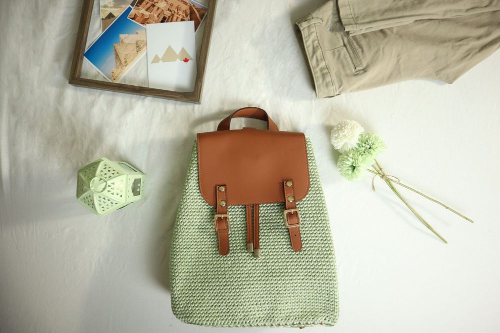 Mochila de crochê verde com detalhes em couro sobre cama branca, entre flores e um quadro, vista de cima