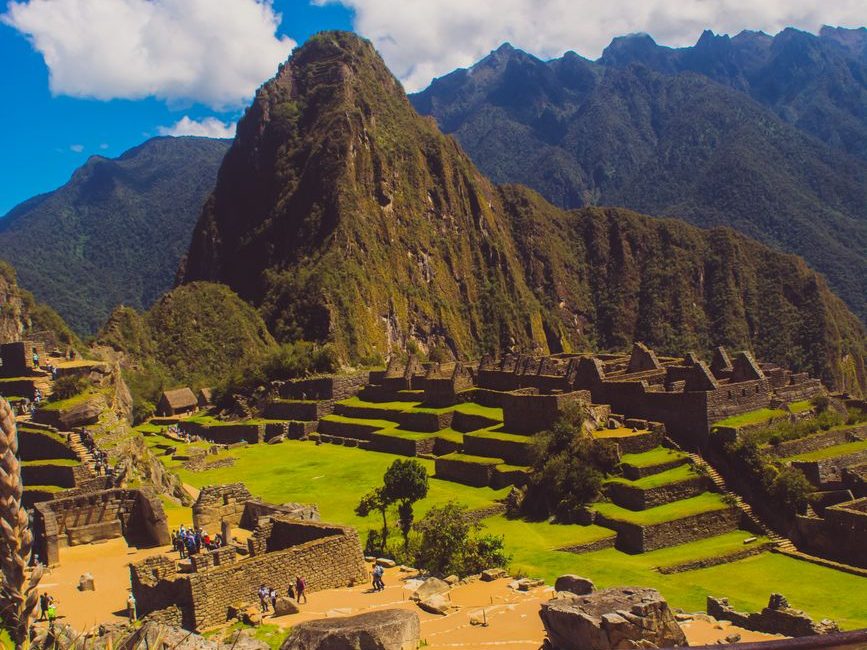 Terraços e construções de pedra de Machu Picchu vistos do alto, com a montanha ao fundo e céu azul