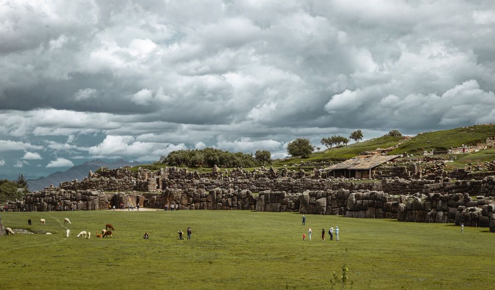 Muralhas de pedra de Sacsayhuamán sobre um gramado verde, com alpacas e céu de nuvens