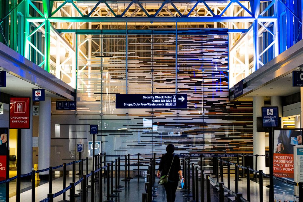 Passageira de costas num corredor de segurança de aeroporto, sob estrutura iluminada em verde e azul