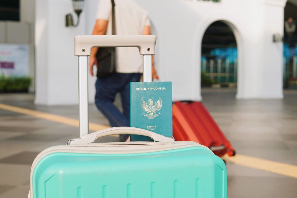 Mala turquesa com um passaporte verde apoiado na alça, em saguão de aeroporto desfocado