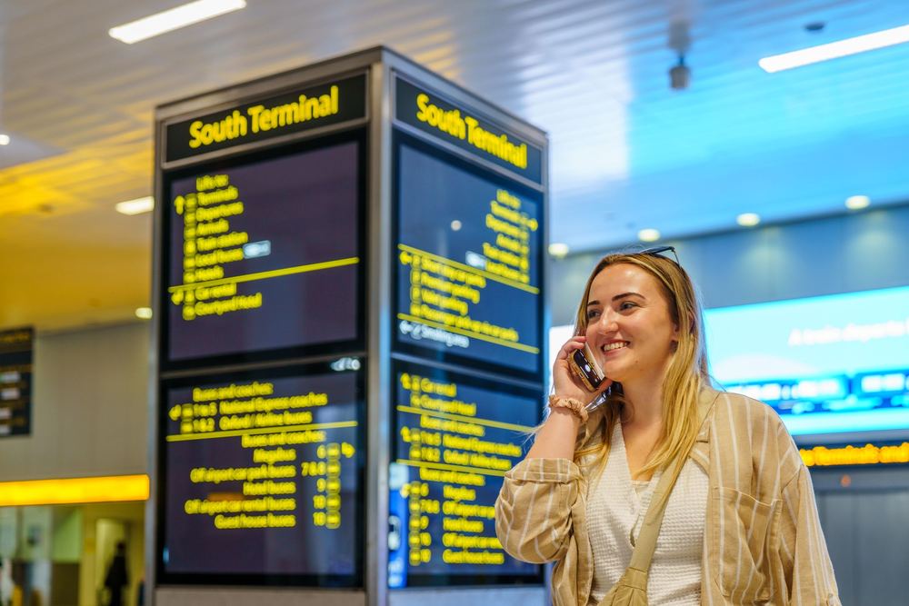 Mulher sorrindo ao telefone ao lado de painéis de informação em terminal de aeroporto