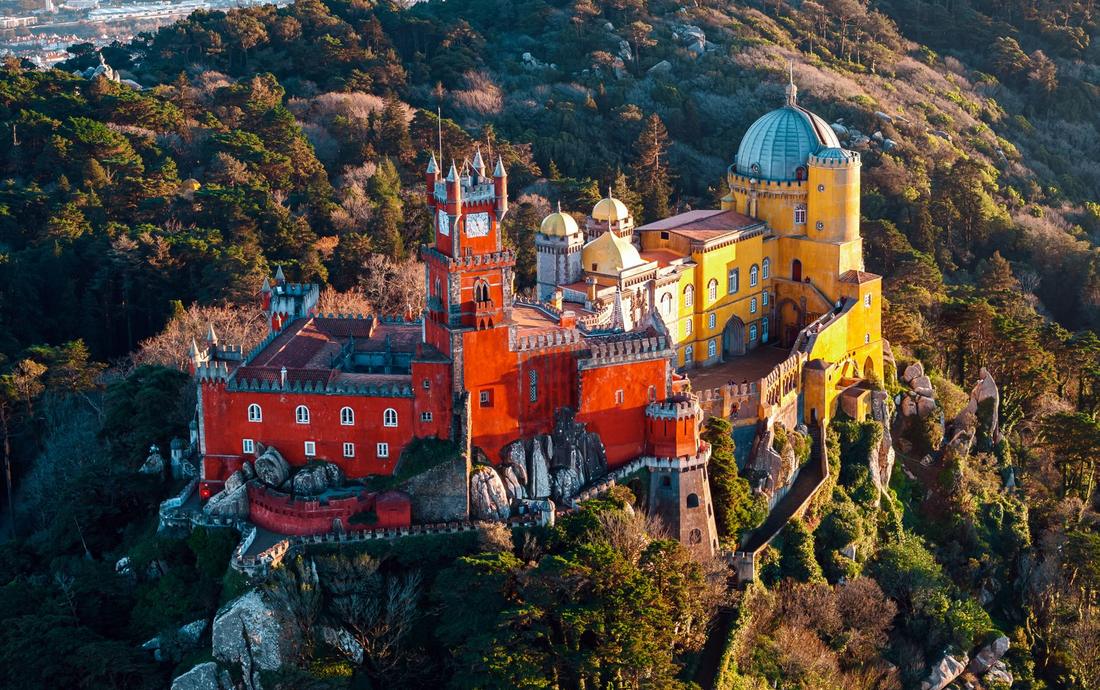Vista aérea do Palácio da Pena, em Sintra, com torres vermelhas e amarelas no alto de um morro coberto de floresta, na luz dourada do fim de tarde