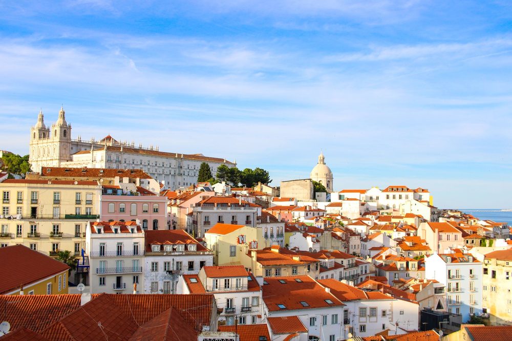Telhados alaranjados e fachadas coloridas de Alfama, com uma igreja branca no alto e céu azul com nuvens