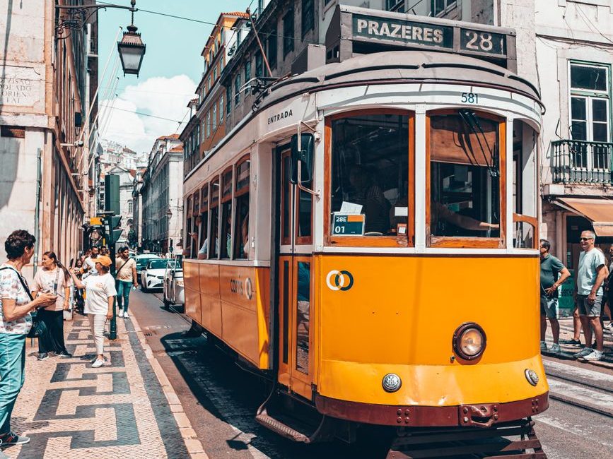 Elétrico amarelo, com a placa Prazeres 28, numa rua de calçada de pedra, entre prédios antigos