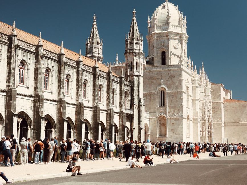 Fachada de pedra clara do Mosteiro dos Jerónimos, com torres rendilhadas, e uma fila de visitantes na calçada