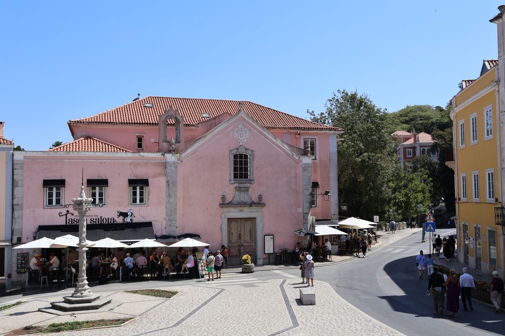 Praça do centro histórico de Sintra, com um casarão rosa de telhado de cerâmica, guarda-sóis brancos e pessoas passeando