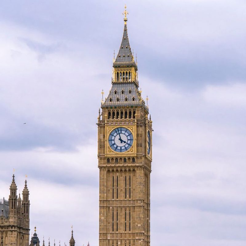 Torre do relógio do Big Ben, com o mostrador azul e dourado, e parte do Parlamento ao lado, sob céu nublado
