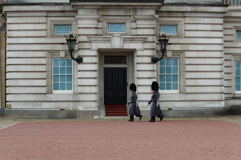 Dois guardas de casaco longo e chapéu de pelo preto caminhando diante de uma porta do Palácio de Buckingham