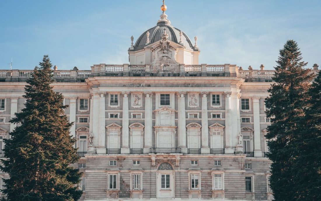 Fachada do Palácio Real de Madri, com a cúpula ao centro e pinheiros dos dois lados, sob um céu azul