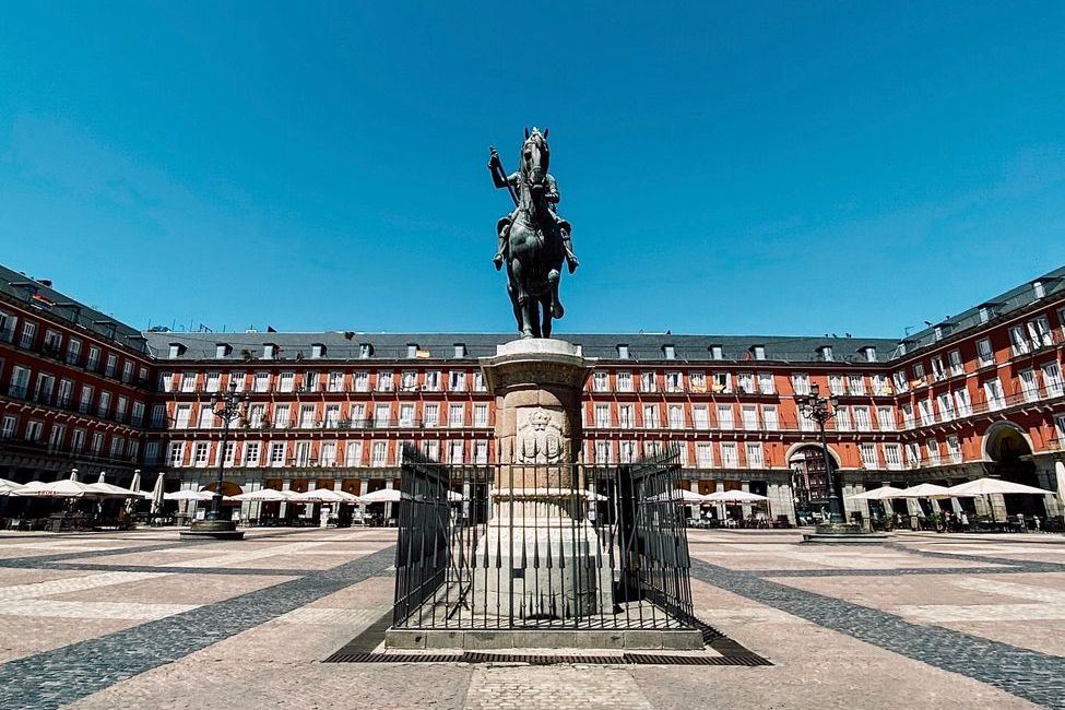 Plaza Mayor de Madri com a estátua equestre ao centro, prédios vermelhos com arcadas ao fundo e céu azul