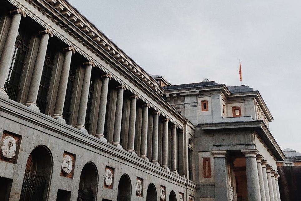 Colunata e fachada de pedra do Museu do Prado, com a bandeira da Espanha no alto, sob um céu nublado