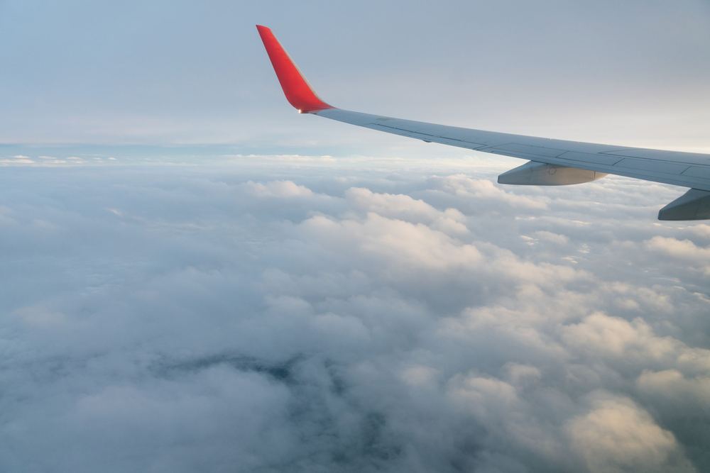 Asa de avião com ponta vermelha sobre um mar de nuvens, em céu claro