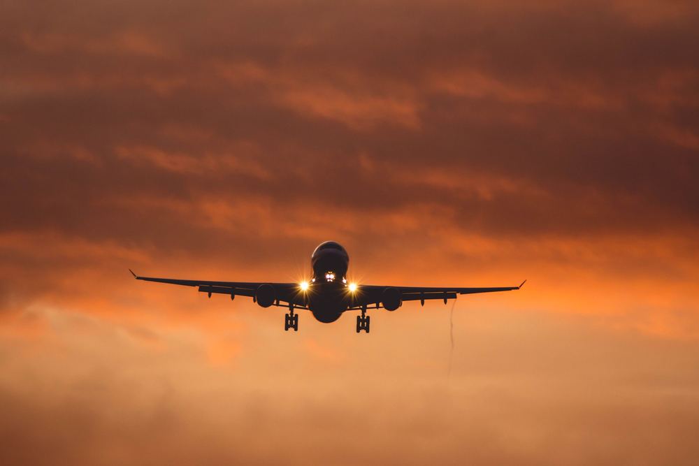 Avião visto de frente em voo, com faróis acesos e céu alaranjado ao entardecer