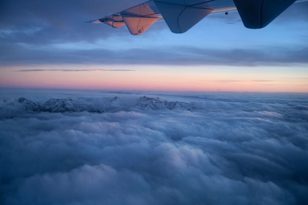 Vista aérea de um mar de nuvens ao entardecer, com céu azul e rosado e ponta de asa no topo