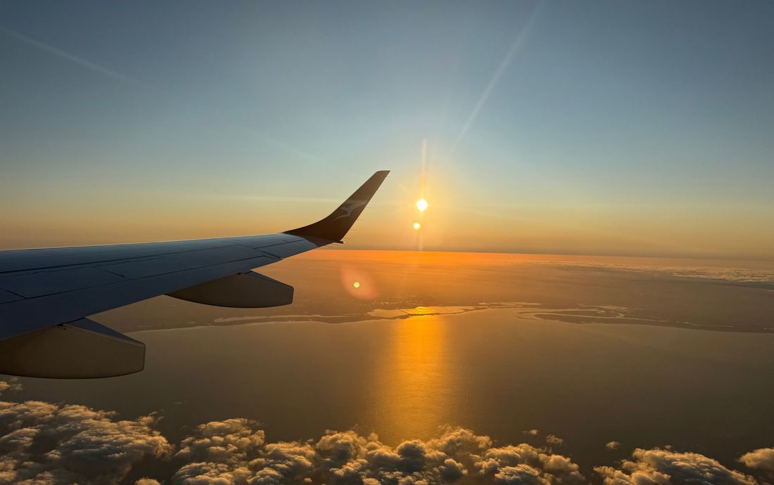 Asa de avião ao pôr do sol, com o sol baixo refletido no mar e nuvens abaixo