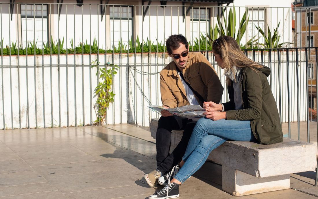 Casal sentado em um banco ao sol, consultando um mapa de papel em um terraço