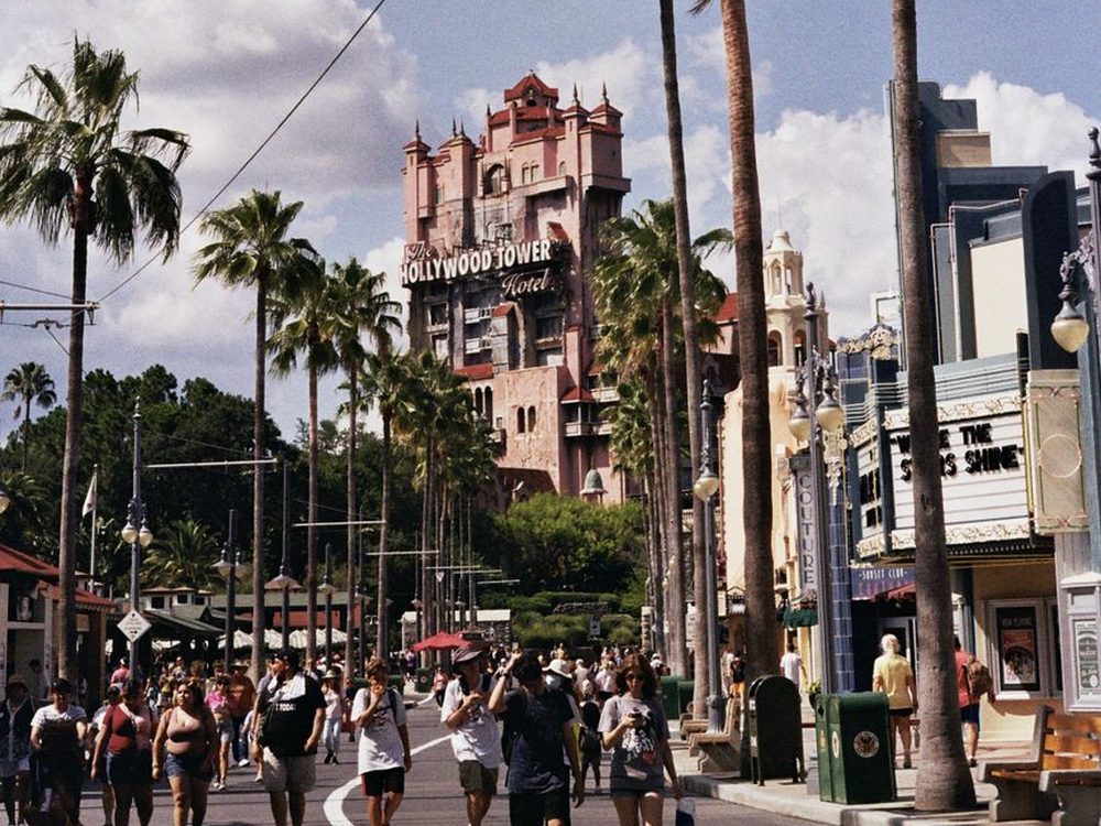 Rua com palmeiras altas e pessoas caminhando, com uma torre rosada ao fundo, no Hollywood Studios