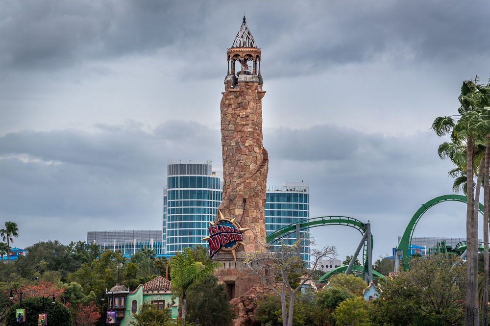 Torre de pedra em forma de farol, com montanha-russa verde e prédios de vidro ao fundo, num dia nublado