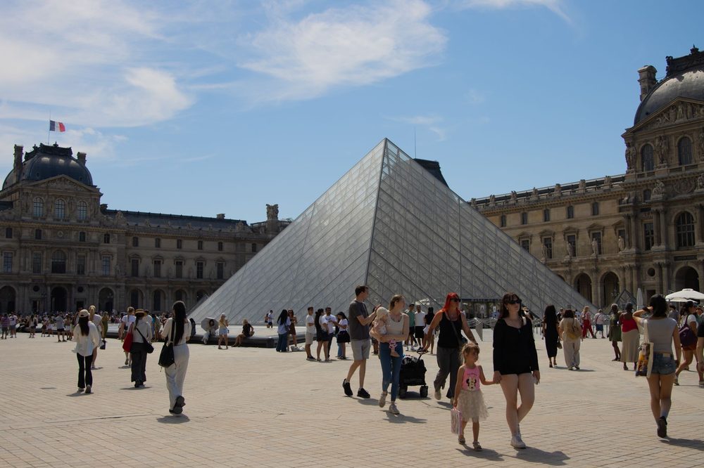 Pirâmide de vidro do Louvre no pátio do museu, com turistas caminhando e o palácio de pedra ao fundo