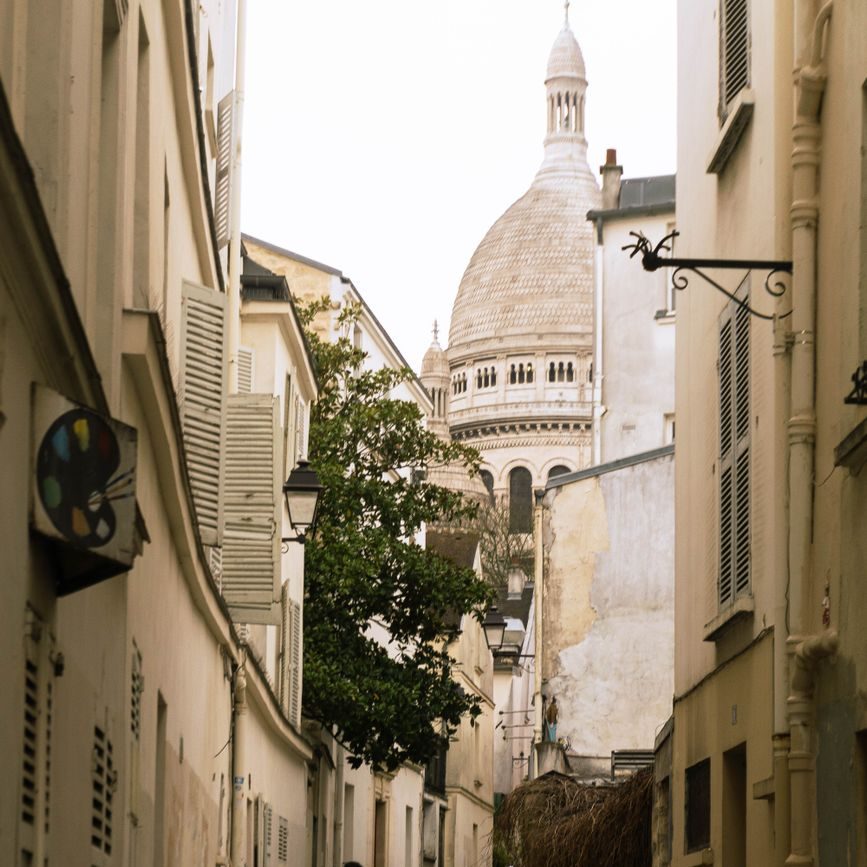 Rua estreita de Montmartre entre casas claras, com a cúpula branca da Basílica de Sacré-Cœur ao fundo
