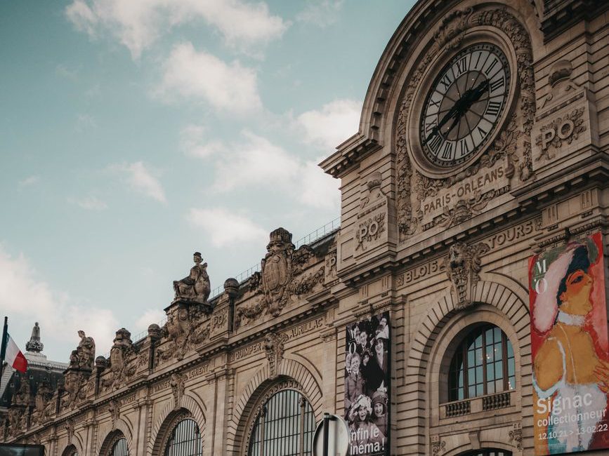 Fachada do Museu d'Orsay, a antiga estação de trem, com o grande relógio de ponteiros pretos no alto e cartazes pendurados nas arcadas