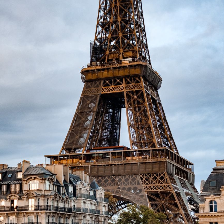 Base e primeiro andar da Torre Eiffel, em ferro marrom-dourado, com prédios de pedra de Paris em primeiro plano e céu nublado