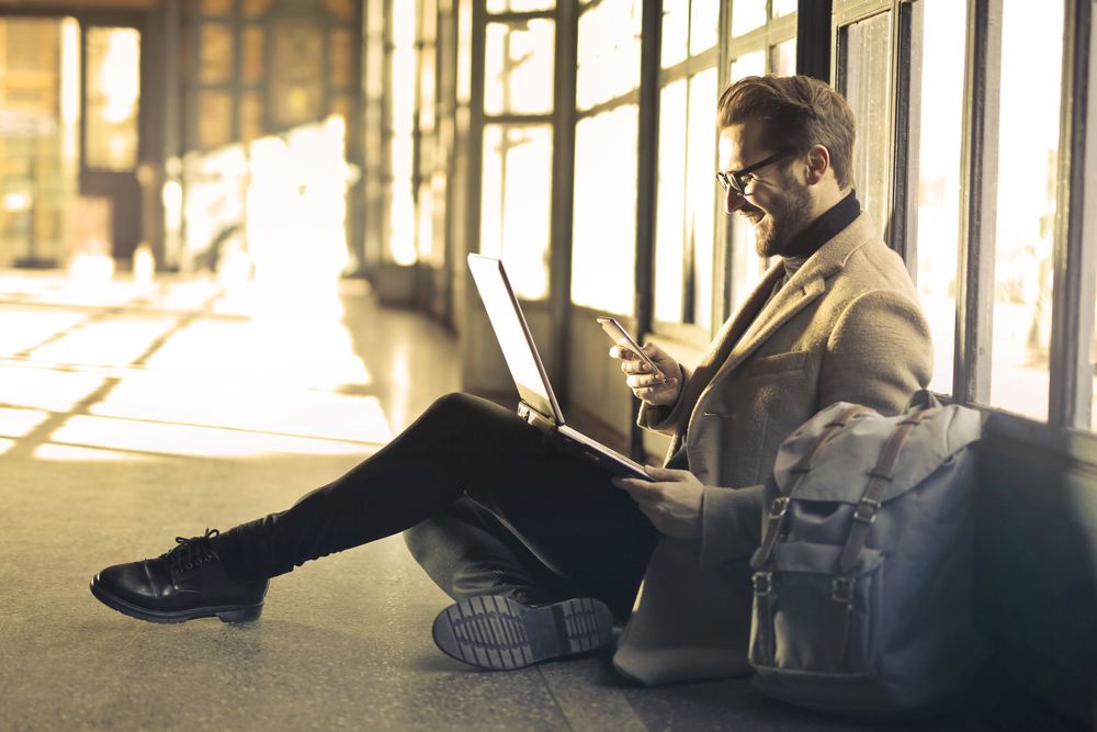 Homem sorrindo com notebook e celular, sentado no chão de um corredor com luz do sol