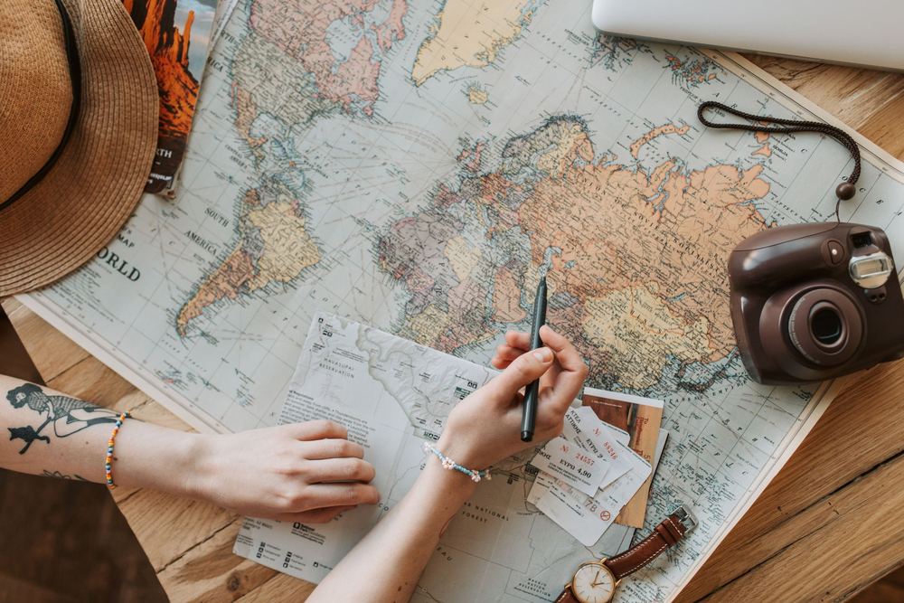 Mãos sobre um mapa do mundo antigo, com chapéu, câmera instantânea e anotações de viagem