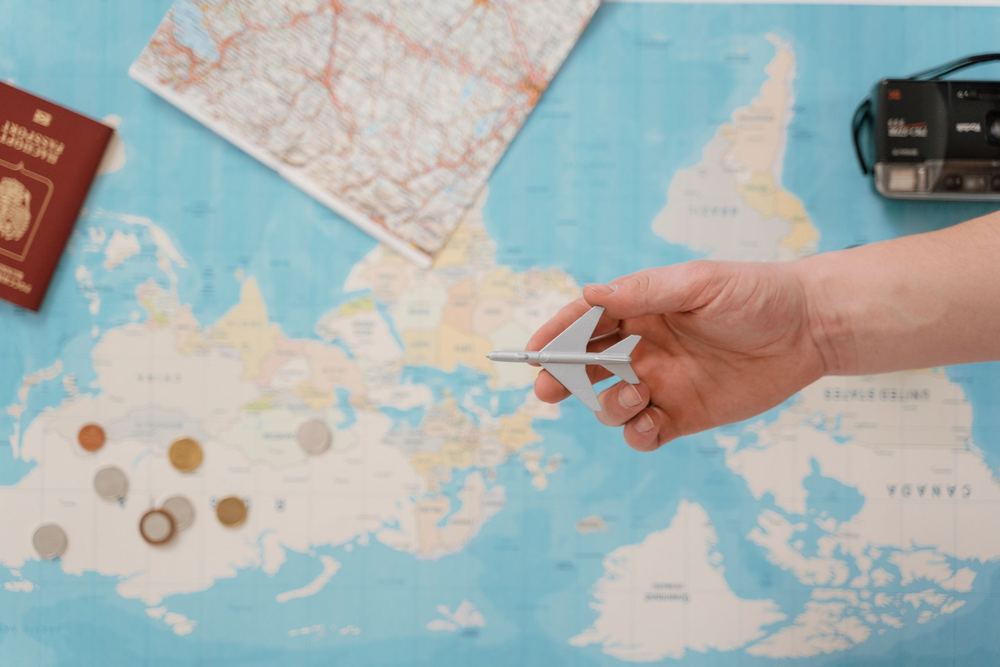 Mão segura um aviãozinho sobre um mapa do mundo azul, com passaporte, câmera e moedas