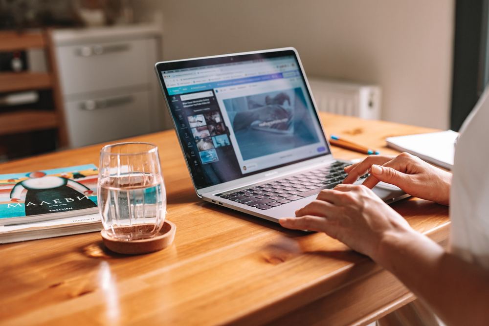 Pessoa digitando em notebook sobre mesa de madeira, com copo de água e revista