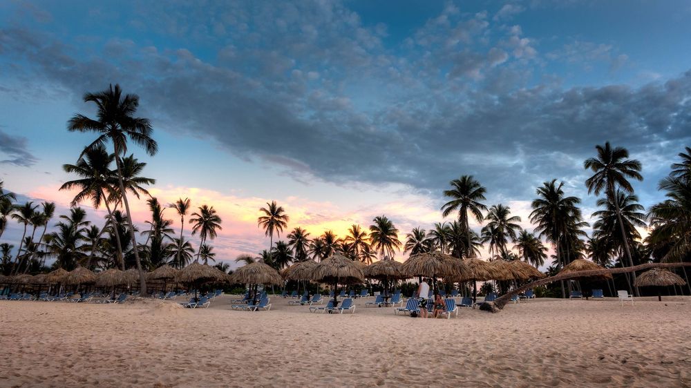 Palmeiras e guarda-sóis de palha numa praia de areia batida, sob um céu de nuvens escuras e um brilho laranja no horizonte