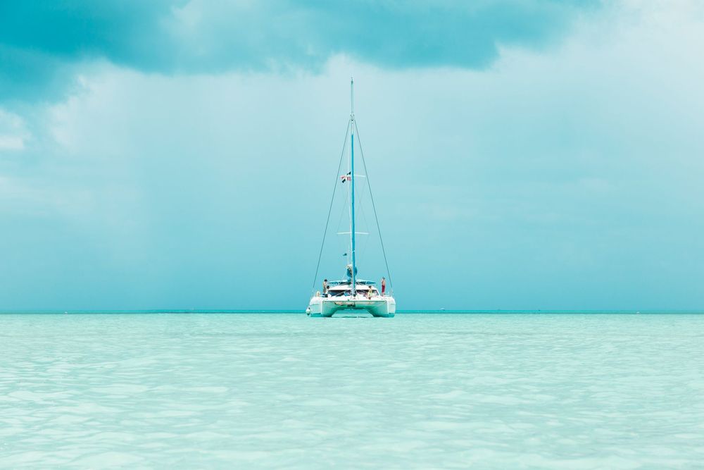 Catamarã de velas recolhidas, ao longe, num mar de águas rasas em tons de turquesa e céu claro
