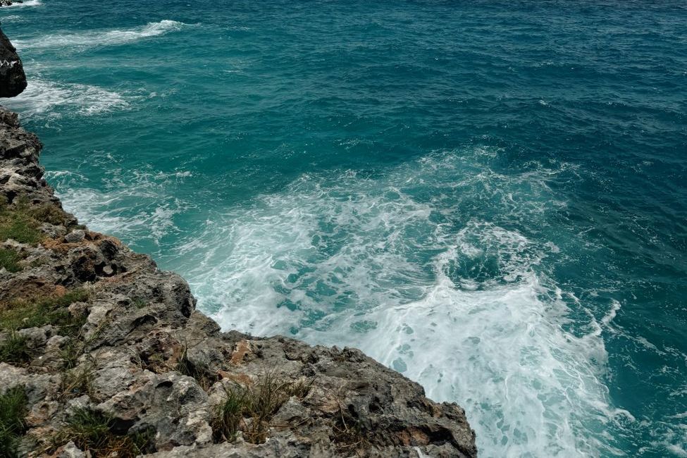 Ondas de espuma branca batendo contra uma costa de pedras, num mar verde-azulado visto do alto