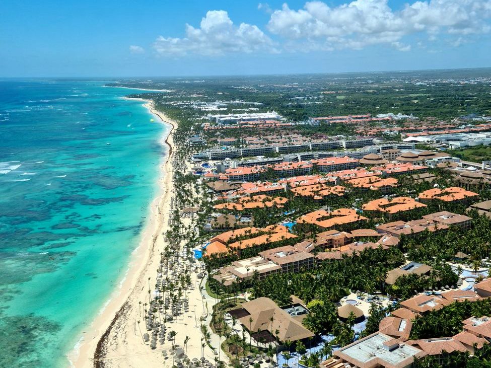 Vista aérea de uma faixa de praia com areia clara, mar turquesa de um lado e hotéis de telhado alaranjado entre árvores do outro