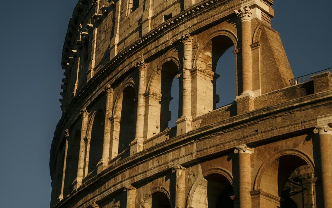 Parede externa do Coliseu de Roma, dourada pela luz do fim de tarde, contra um céu azul-escuro