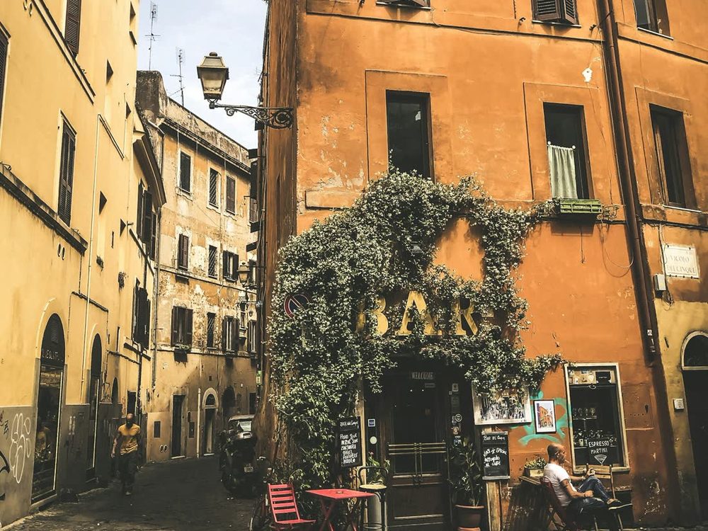 Rua de pedras do Trastevere, com casas em tons de ocre, trepadeiras e mesas de um bar na calçada