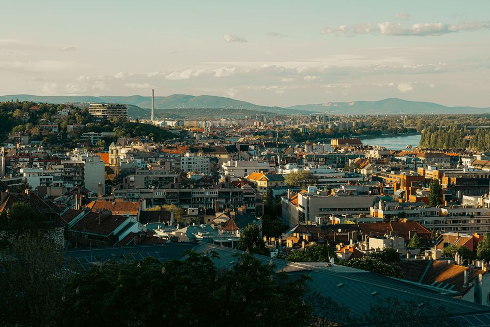Vista do alto de uma cidade com telhados, rio e colinas ao fundo, em luz de fim de tarde