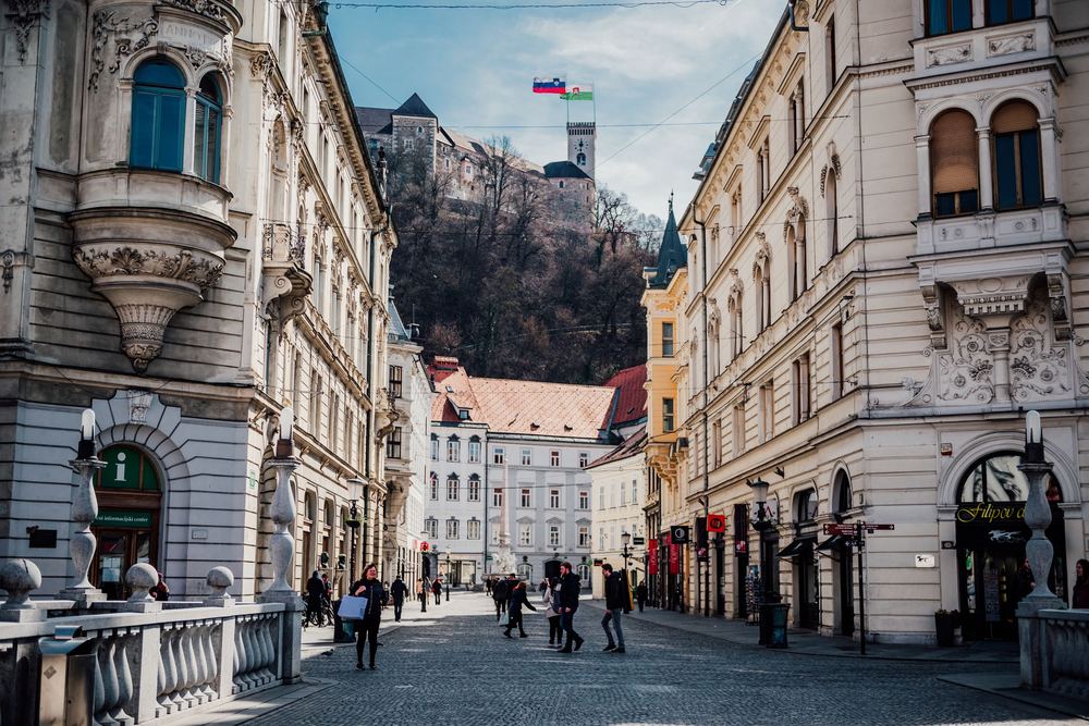 Rua de pedestres com prédios históricos, castelo em uma colina e céu azul ao fundo
