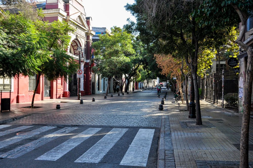 Rua arborizada do Barrio Bellavista, em Santiago, com faixa de pedestres e um prédio vermelho ao fundo